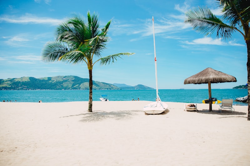 White sand beach with a palm tree, a beached sailboat, and a thatched umbrella against green mountains